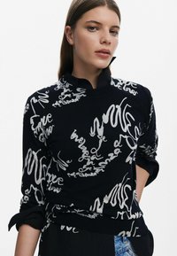 Svart tröja med vita handskrivna mönster, med en mockkrage och upprullade ärmar. Mjuk textur och figurnära design.
