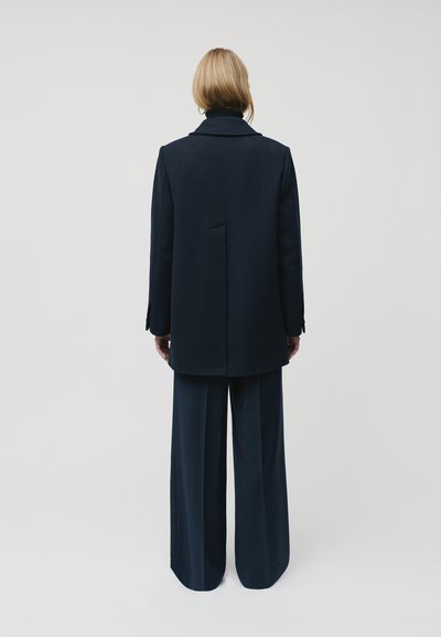 Navy oversized mētelis ar izgrieztu apkakli, aizmugures venti, un garām piedurknēm, pārī ar platām biksēm tajā pašā krāsā, rādīts no aizmugures.
