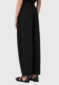 Pantalones anchos negros de talle alto con una textura suave. Presentan un lazo para cinturón y detalles laterales. Combinados con zapatos planos negros.