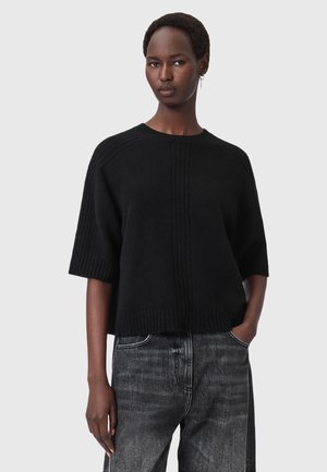 Zwarte gebreide pullover met korte mouwen, geribbelde textuur en verticale naaddetail. Gecombineerd met grijze spijkerbroek voor contrast.