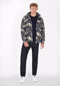 Giacca camouflage con cappuccio nei toni grigio, blu navy e crema; abbinata a jeans scuri e sneakers bianche. Texture liscia e design casual.