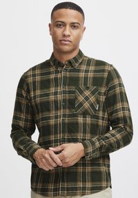 Camisa de franela a cuadros verde y marrón con cuello abotonado, mangas largas y un bolsillo en el pecho. Textura suave y diseño de corte regular.