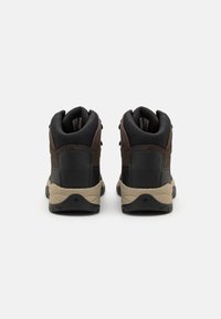 Pár hnědých a černých turistických bot zobrazených ze zadní strany, s polstrovanými límci a béžovými textured podrážkami na bílém pozadí.