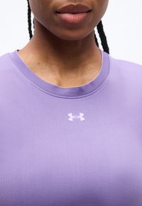 Κοντινό πλάνο ενός ατόμου που φοράει μοβ μπλούζα με στρογγυλή λαιμόκοψη, όπου υπάρχει ένα μικρό λευκό λογότυπο Under Armour στο κέντρο κοντά στον γιακά.