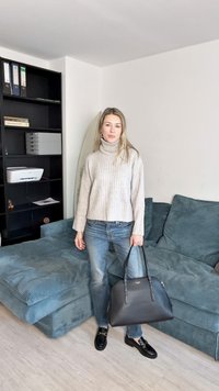 Világosszürke bordázott garbó, kék farmernadrág, fekete kézitáska és fekete loaferek. A háttérben kanapé és könyvespolc, irodai kellékekkel.