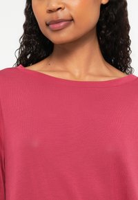 Pink lightweight top with a wide neckline and textured fabric, featuring short sleeves and a relaxed fit. Close-up view of the garment.