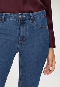 Ciemnoniebieskie dżinsy mają wysoki stan, pięć kieszeni, dekoracyjne przeszycia oraz srebrny guzik z przodu.