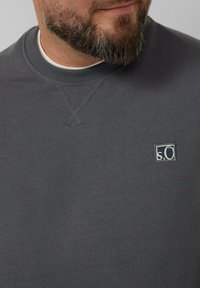 Primo piano del volto inferiore di un uomo con una barba curata, che indossa una felpa grigia a girocollo con un piccolo logo "s.O".