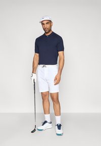 Ναυτικό πόλο μπλούζα, άσπρα σορτς, άσπρα παπούτσια με μπλε λεπτομέρειες, κρατώντας ένα γκολφ κλαμπ. Αξεσουάρ περιλαμβάνουν ένα άσπρο καπέλο και ένα γάντι.