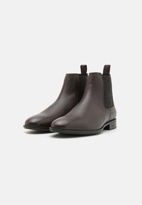 Bottes Chelsea en cuir marron avec une finition lisse, panneaux latéraux élastiques, languettes de tirage et semelles plates, arborant un design minimaliste et sans motifs visibles.