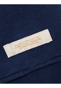 Marineblauwe stof met een gestructureerde brei. Heeft een crèmekleurig label dat aan de onderkant is gestikt, met gouden tekst waarop merkinformatie staat.