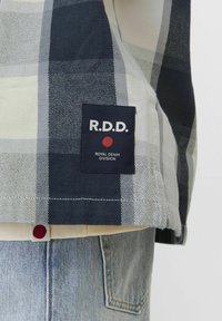 Camicia a scacchi blu e bianca in cotone con etichetta navy che presenta "R.D.D." e un punto rosso, orlata in fondo con cuciture decorative.