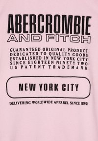 Lys rosa hettegenser med svart tekst. Designet inkluderer "ABERCROMBIE AND FITCH" øverst, med "NEW YORK CITY" uthevet i fet skrift.