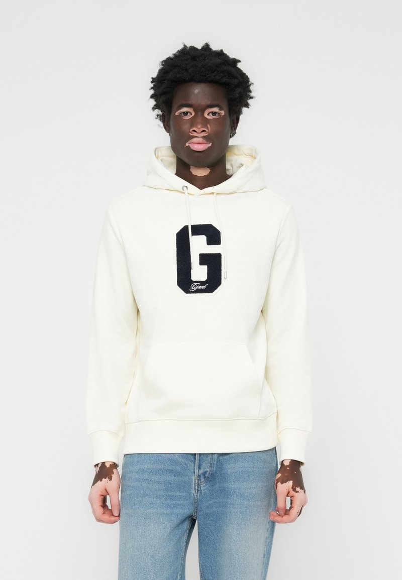 Sudadera crema con un gran logo bordado en navy de la "G" en el pecho, con un bolsillo frontal y capucha con cordón, combinada con jeans de color azul claro.