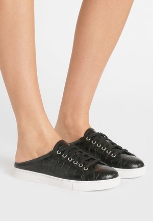 Zwarte instapsneakers met krokodillenstructuur, zwarte veters en witte rubberen zolen, gedragen op blote voeten tegen een lichte achtergrond.
