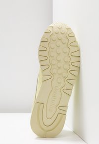 Semelle d'une chaussure Reebok beige clair montrant le motif de la bande de roulement et le logo, positionnée verticalement contre un mur d'angle blanc.