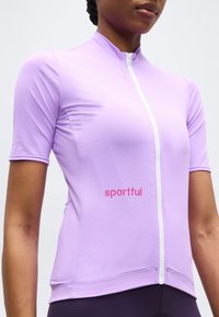 Γυναίκα που φοράει εφαρμοστή κοντομάνικη ποδηλατική φανέλα σε ανοιχτό μωβ χρώμα με λευκό φερμουάρ μπροστά και το λογότυπο "sportful" σε κόκκινο στο στήθος.