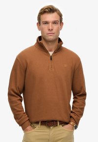Brauner, mit Reißverschluss versehenen Pullover mit langen Ärmeln, geripptem Muster und kleinem Logo auf der Brust, kombiniert mit beigen Hosen und einem geflochtenen Gürtel.