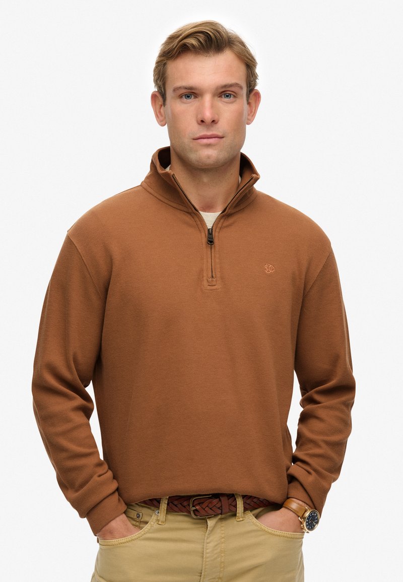 Brauner, mit Reißverschluss versehenen Pullover mit langen Ärmeln, geripptem Muster und kleinem Logo auf der Brust, kombiniert mit beigen Hosen und einem geflochtenen Gürtel.