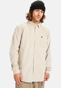 Camisa de tergal beige con diseño de botones en el frente, mangas largas y un bolsillo en el lado izquierdo del pecho, combinada con un gorro de punto negro.
