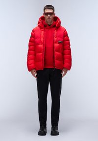 Giacca rossa puffa con cappuccio, dotata di tasche con zip e patch del logo. Indossata sopra un hoodie rosso coordinato e abbinata a pantaloni neri e stivali.