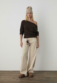 Brauner schulterfreier Pullover, beigefarbene weit geschnittene Hose und braune Slipper. Schlüsselanhänger mit einem kleinen Spielzeug an der Hose. Neutraler Hintergrund.