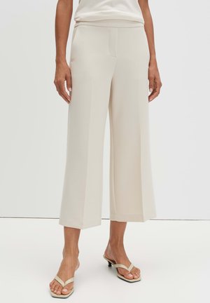 Persona che indossa pantaloni cropped a gamba larga color crema e sandali beige con tacco, in piedi su uno sfondo chiaro e semplice.