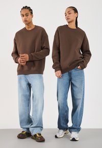 To personer iført brune hettegensere og løse lyseblå jeans står mot en ensfarget bakgrunn, vendt framover.
