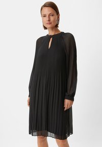 Schwarzes, plissiertes Kleid mit langen, durchsichtigen Ärmeln, Schlüssel-ausschnitt und gerafften Manschetten. Der Stoff hat eine weiche Textur und eine fließende Silhouette.