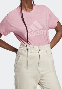 Vaaleanpunainen pilkullinen puuvillainen t-paita, jossa valkoinen Adidas-logo, yhdistetty korkeavyötäröisiin off-white-housuihin, joissa on napit ja taskut.