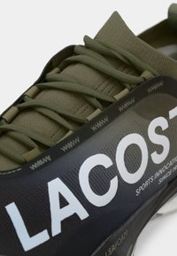 Lacoste sportska obuća s tamnozelenim gornjim dijelom, teksturiranim pletenim materijalom, istaknutim logotipom i fleksibilnim vezicama. Ležeran dizajn s detaljima odvojene podloge.
