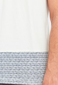 Kortærmet t-shirt i hvid bomuld med et horisontalt marineblåt stribet panel i kanten, der har en glat tekstur og kontrasterende mønstre.