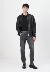 Jachetă bomber din piele neagră cu închidere cu fermoar, blugi gri cu o ușoară decolorare și cizme negre până la gleznă. Modelul stă în fața unui fundal simplu.