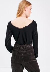 Maglietta nera a maniche lunghe con scollo a V e bordi in pizzo, con dettagli in rete sulle maniche, abbinata a pantaloni di velluto a coste scuri con tasche.