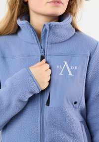 Giacca in pile di colore azzurro chiaro con chiusura a zip, collo alto e tasca sul petto con logo ricamato. Tessuto strutturato con finitura morbida.