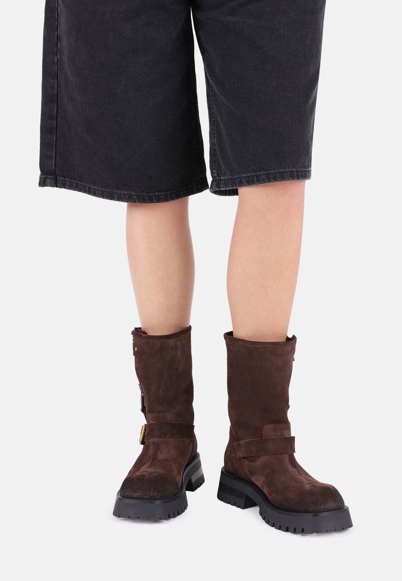 Braune Wildleder-Ankle-Boots mit strukturierter Oberfläche, dicker schwarzer Gummisohle und Riemen-Detail um den Knöchel, kombiniert mit schwarzen Denim-Shorts.
