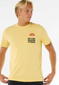 Gul t-shirt lavet af bomuld, med en frontlomme og et grafisk motiv i sort og orange tekst, der lyder "RIP CURL SOAKING UP THE SUN."