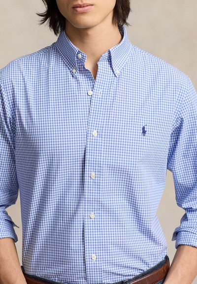 Mees, kes kannab helesinist ja valget ruudulist nööpidega särki, mille varrukad on üles keeratud, ning tumeid teksapükse, seisab neutraalse tausta ees.