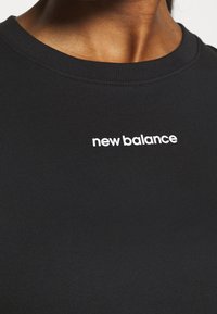 Camiseta negra confeccionada en tela suave y elástica con cuello redondo, que presenta un logo blanco de "new balance" en el centro.