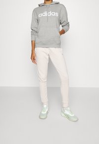 Felpa grigia con logo "adidas" bianco, tasca anteriore, abbinata a pantaloni jogger rosa chiaro e sneakers verde menta con strisce bianche.