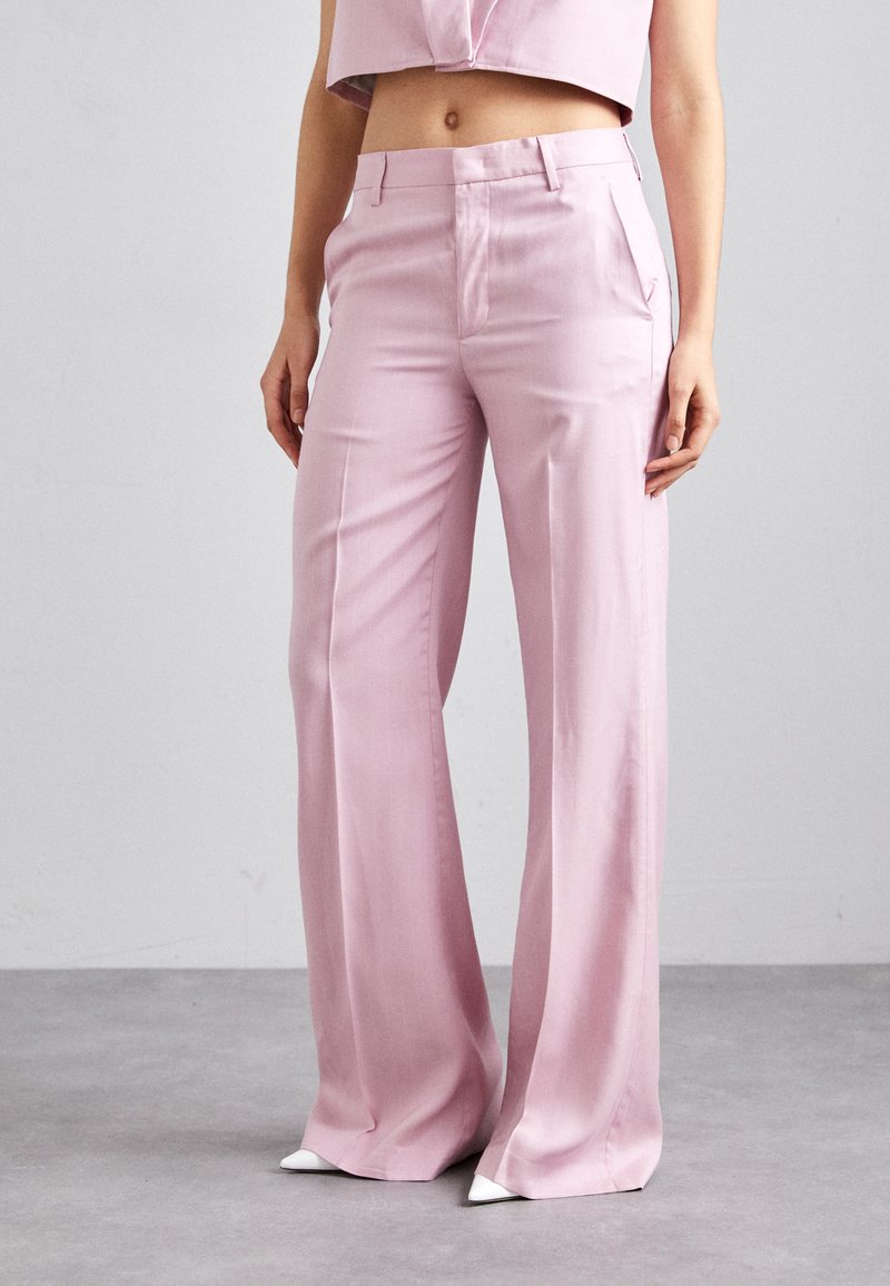 Pantalons larges rose clair en tissu lisse, dotés d'une taille haute et de poches avant, associés à des chaussures blanches à bout pointu.