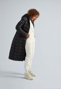 Lang sort pufferjakke med hætte, der har et quiltet design. Parret med en hvid sweater og hvide joggere. Lyse sneakers.
