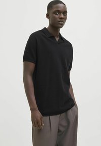 Polo shirt nera a maniche corte con un tessuto a maglia testurizzato, caratterizzata da un collo a V e una vestibilità comoda. Indossata con pantaloni grigi chiari su misura.