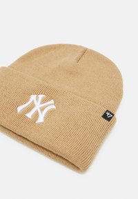 Gorro de punto marrón con vuelta, logotipo "NY" bordado en blanco y una pequeña etiqueta negra con texto blanco. Tela suave y texturizada.