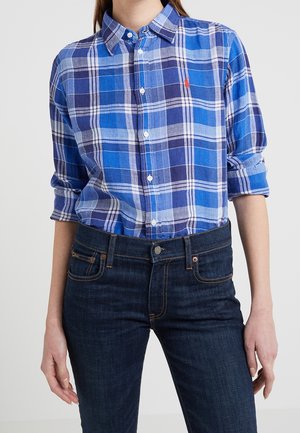 Woman wearing blue and white plaid button-up shirt with rolled sleeves tucked into dark blue jeans.