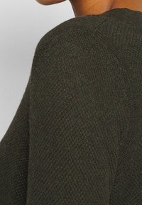 Maglione lavorato a maglia verde scuro con una finitura testurizzata e colletto a coste, che mostra la spalla e la parte superiore del braccio in una vista ravvicinata.