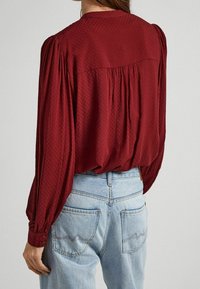 Röd texturerad blus med lång ärm, rynkade axelsömmar och knappmanchetter, kombinerad med ljusblå denimjeans med böjd fickstickning.