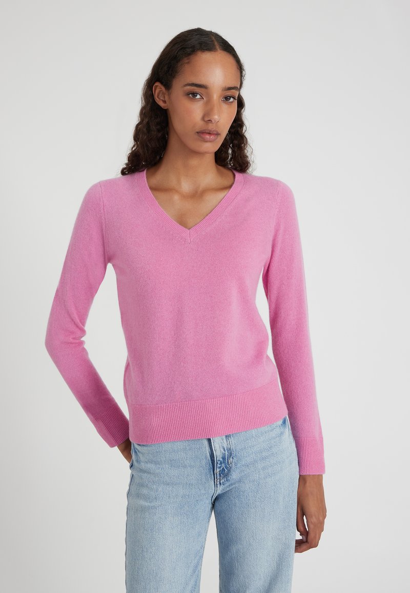 Donna con lunghi capelli ricci, che indossa un maglione rosa a scollo a V e jeans azzurri chiari, in piedi su uno sfondo grigio semplice.
