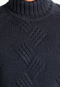 Dunkelblauer gestrickter Pullover mit hohem Rippkragen. Verfügt über ein strukturiertes Zopfmuster, das dickes Garn und dimensionale Linien zur Geltung bringt.