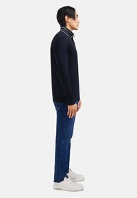 Homme de profil portant un pull bleu marine avec un col rayé, un jean bleu et des baskets blanches sur fond clair.